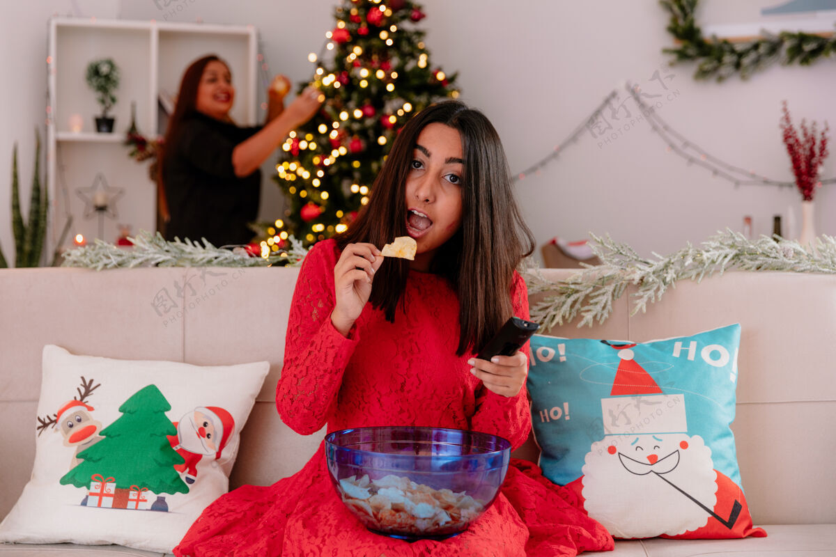 薯条微笑的母亲装饰圣诞树 看着女儿坐在沙发上吃着一碗薯条 在家里享受圣诞节时光微笑坐圣诞快乐