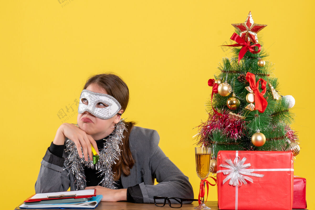 圣诞节前景色漫步的年轻女孩戴着化装面具坐在圣诞树和鸡尾酒礼物的桌边礼物肖像喜剧演员