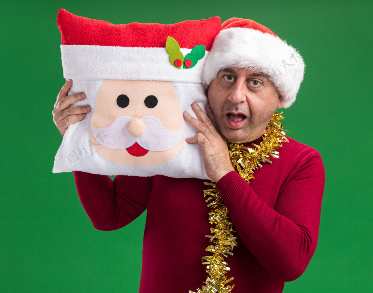 枕头中年男子戴着圣诞老人帽 脖子上戴着金属丝 抱着圣诞枕头 站在绿色的背景下惊讶地看着相机惊讶背景金属片