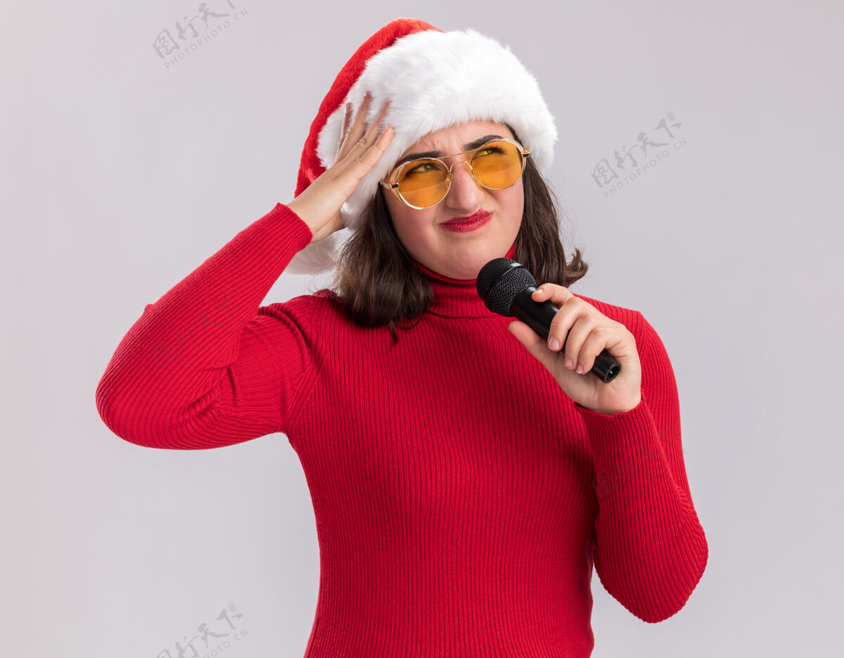 圣诞老人穿着红毛衣 戴着圣诞帽 戴着眼镜 拿着麦克风的年轻女孩站在白色的墙上 困惑而不高兴地看着一边毛衣麦克风站着