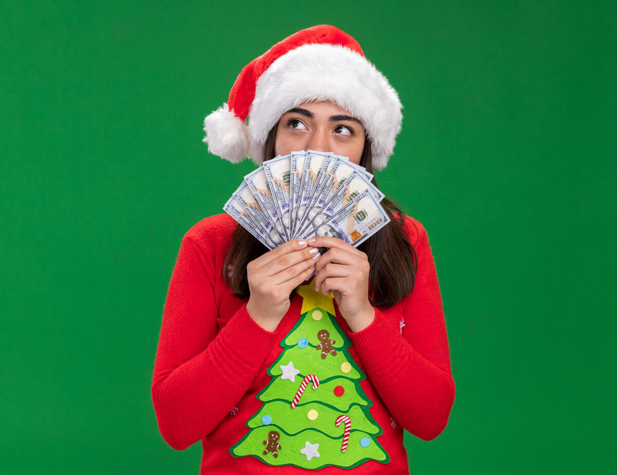 年轻高兴的年轻白人女孩戴着圣诞帽拿着钱看着一边孤立的绿色背景与复制空间圣诞快乐看圣诞节
