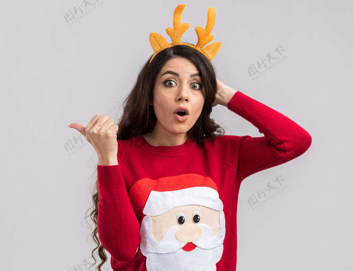 头惊讶的年轻漂亮女孩戴着驯鹿鹿角头带和圣诞老人毛衣看着手放在头上指着侧面鹿角毛衣新