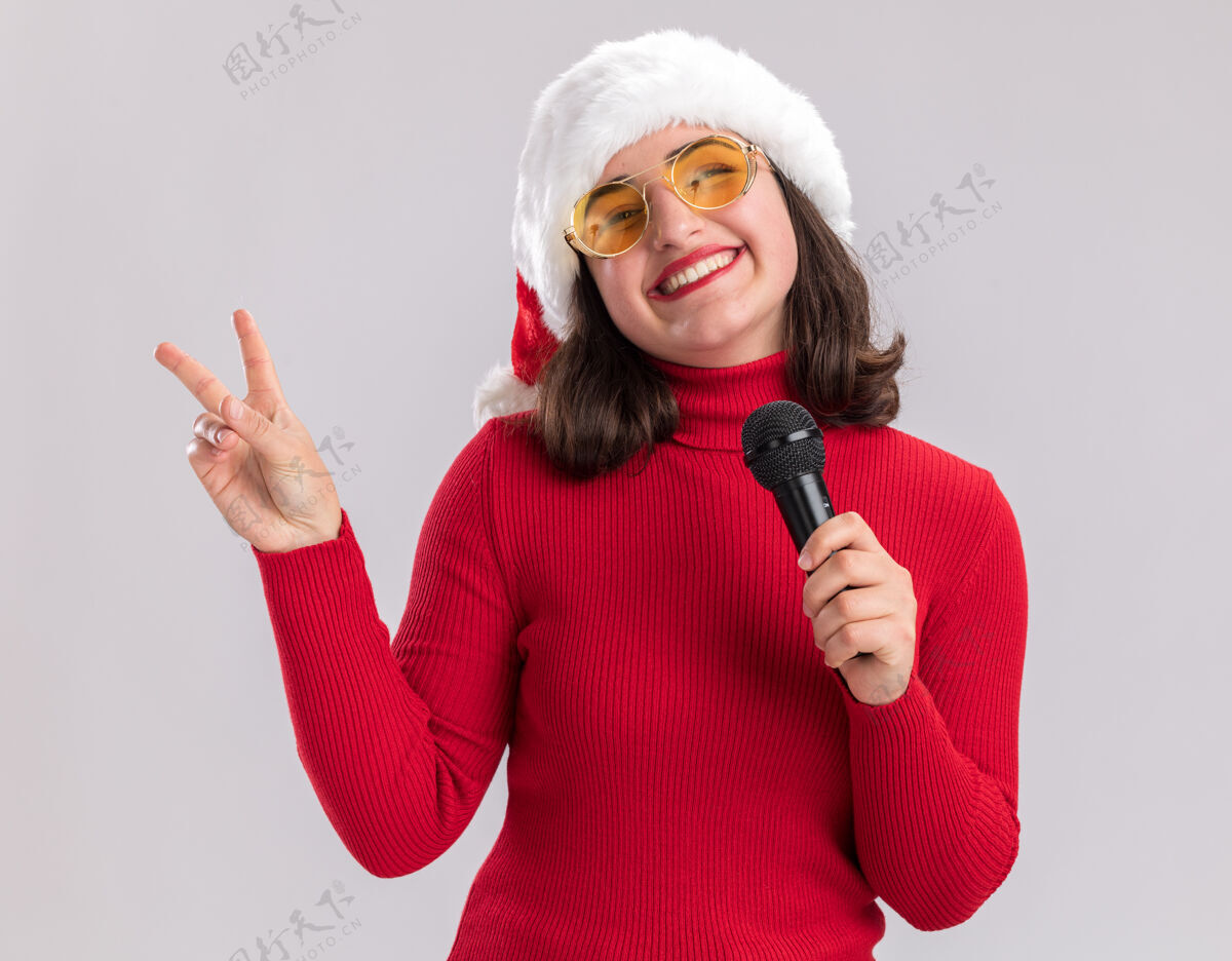 圣诞节快乐快乐的年轻女孩 穿着红色毛衣 戴着圣诞帽 戴着眼镜 拿着麦克风 兴高采烈地微笑着 白色的墙上挂着v字标志毛衣圣诞老人穿着
