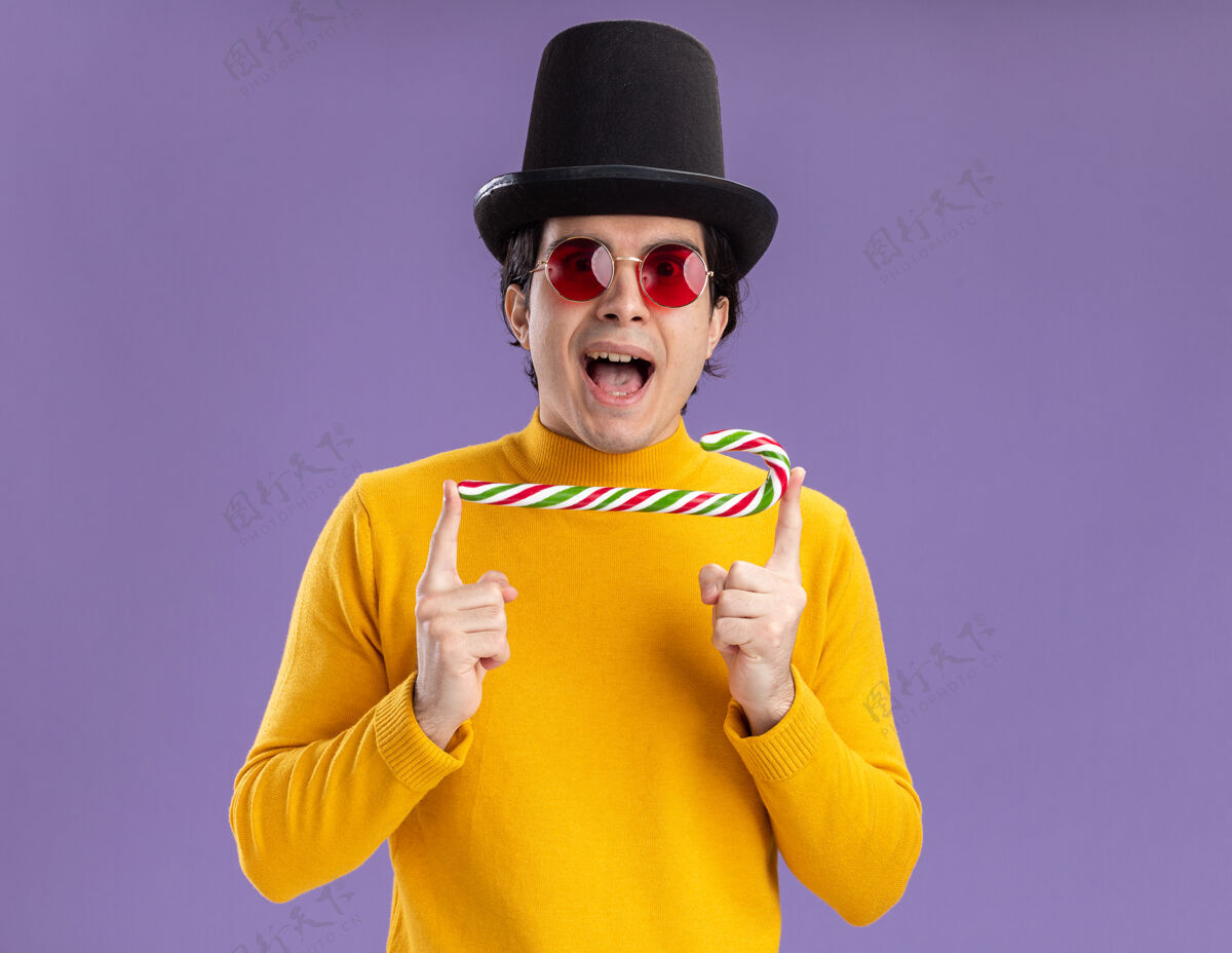 站着穿着黄色高领毛衣戴着眼镜戴着黑帽子拿着糖果手杖快乐地站在紫色的墙上的年轻人高兴地拐杖高领毛衣