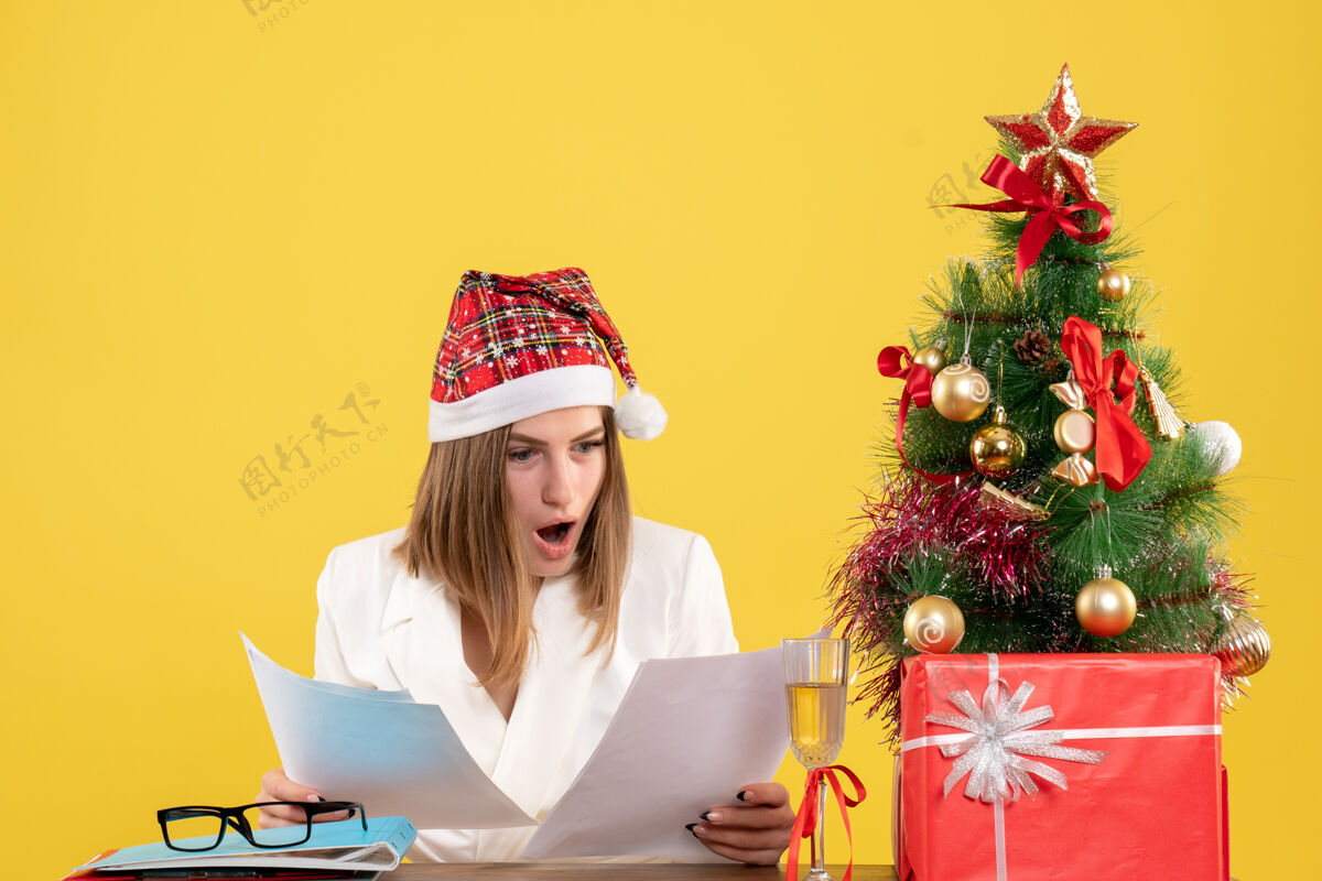 健康正面图：女医生坐在黄色的桌子上 手里拿着圣诞礼物 手里拿着文件微笑快乐漂亮