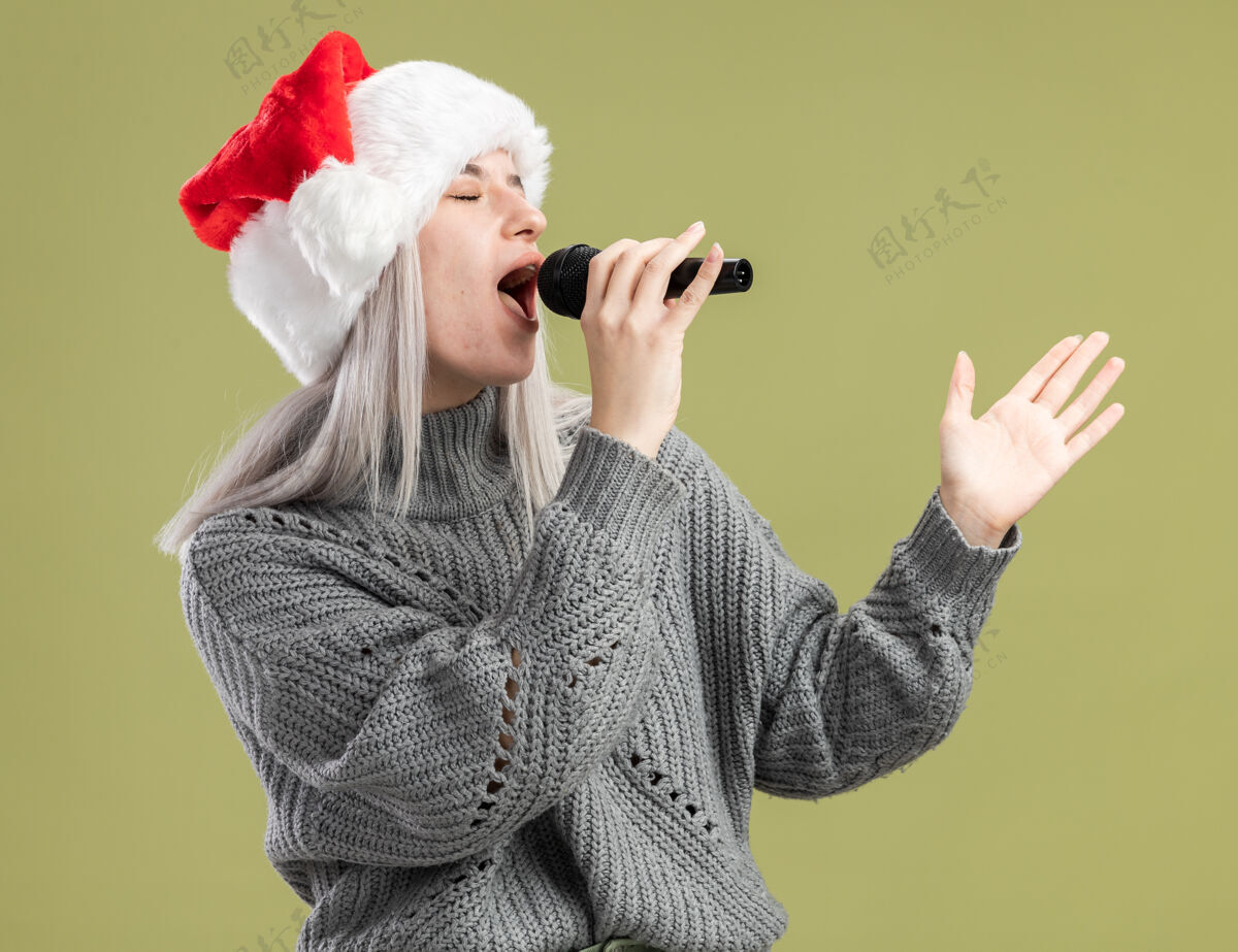 女人年轻的金发女郎穿着冬衣 戴着圣诞帽 手持麦克风 快乐而积极地庆祝圣诞晚会唱歌金发圣诞
