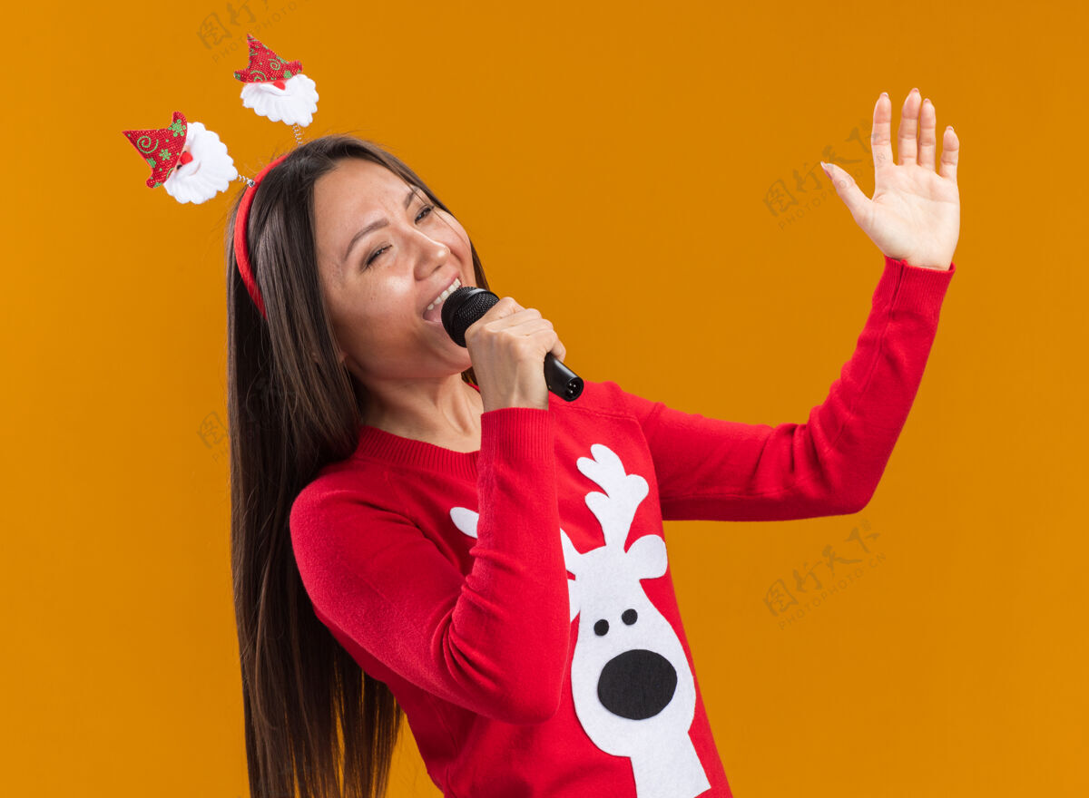 毛衣快乐的年轻亚洲女孩戴着圣诞发箍 穿着毛衣 在橙色墙上的麦克风上唱歌年轻快乐穿着