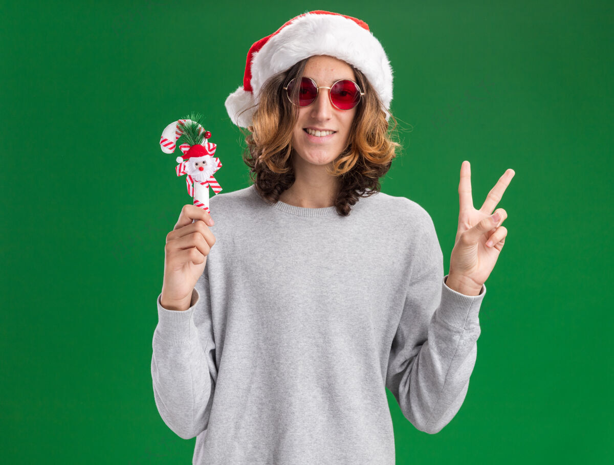 高兴一个戴着圣诞老人帽 戴着红色眼镜 手持圣诞糖果手杖的快乐男人看着相机 愉快地微笑着 在绿色背景上展示着v形标志圣诞老人相机快乐