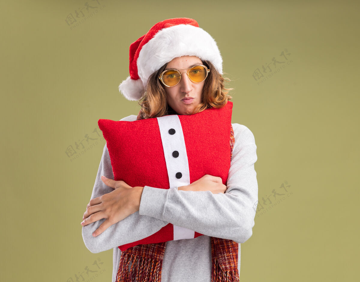 快乐愤愤不平的年轻人戴着圣诞老人的帽子 戴着黄色的眼镜 脖子上围着暖和的围巾 抱着圣诞枕头 站在绿色的背景上 看着摄像机噘着嘴唇圣诞快乐相机怨恨