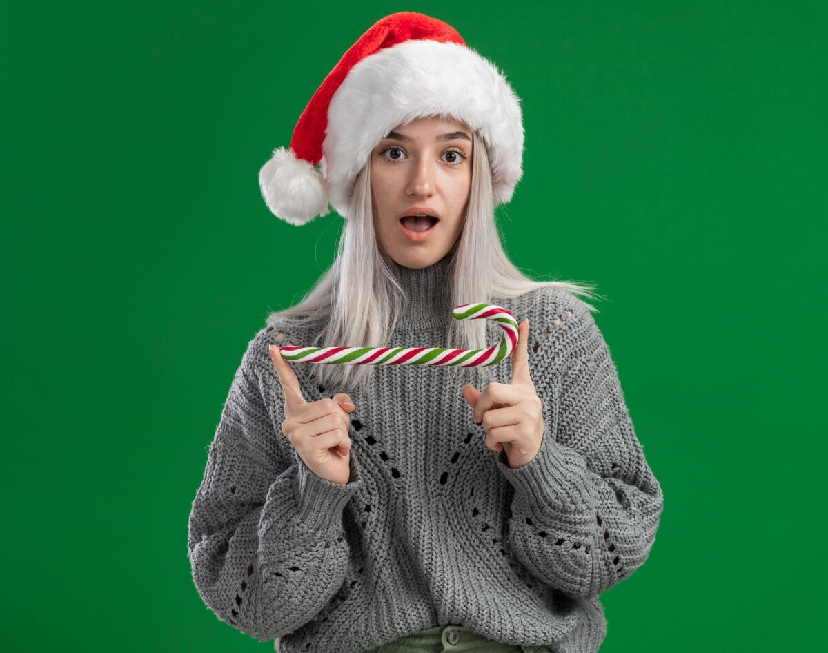 手杖年轻的金发女郎穿着冬天的毛衣 戴着圣诞老人的帽子 手里拿着糖果手杖 站在绿色的背景下惊讶地看着镜头帽子圣诞老人拿着