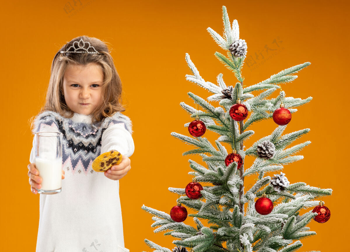 饼干皱着眉头的小女孩站在圣诞树旁 戴着头饰 脖子上戴着花环 手里拿着一杯牛奶 橘黄色的墙上隔着饼干戴着站着在附近