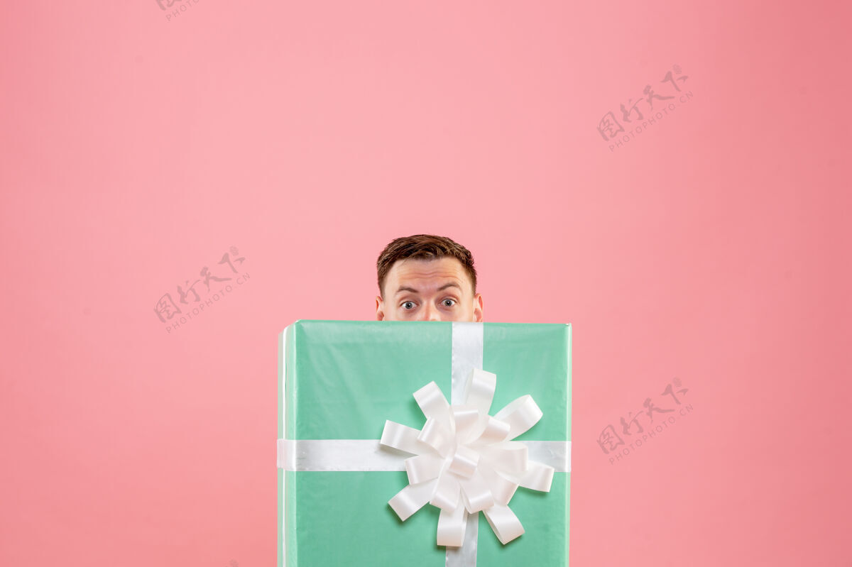 前面隐藏在粉红色墙上的礼物里面的年轻人的正面视图圣诞快乐丝带圣诞节