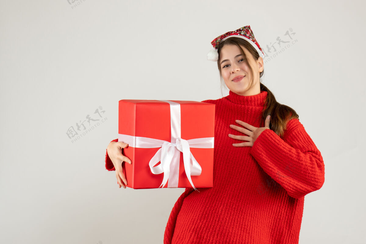 抱着戴着圣诞帽的快乐女孩拿着礼物把手放在胸前站在白色的地板上胸前女孩站着