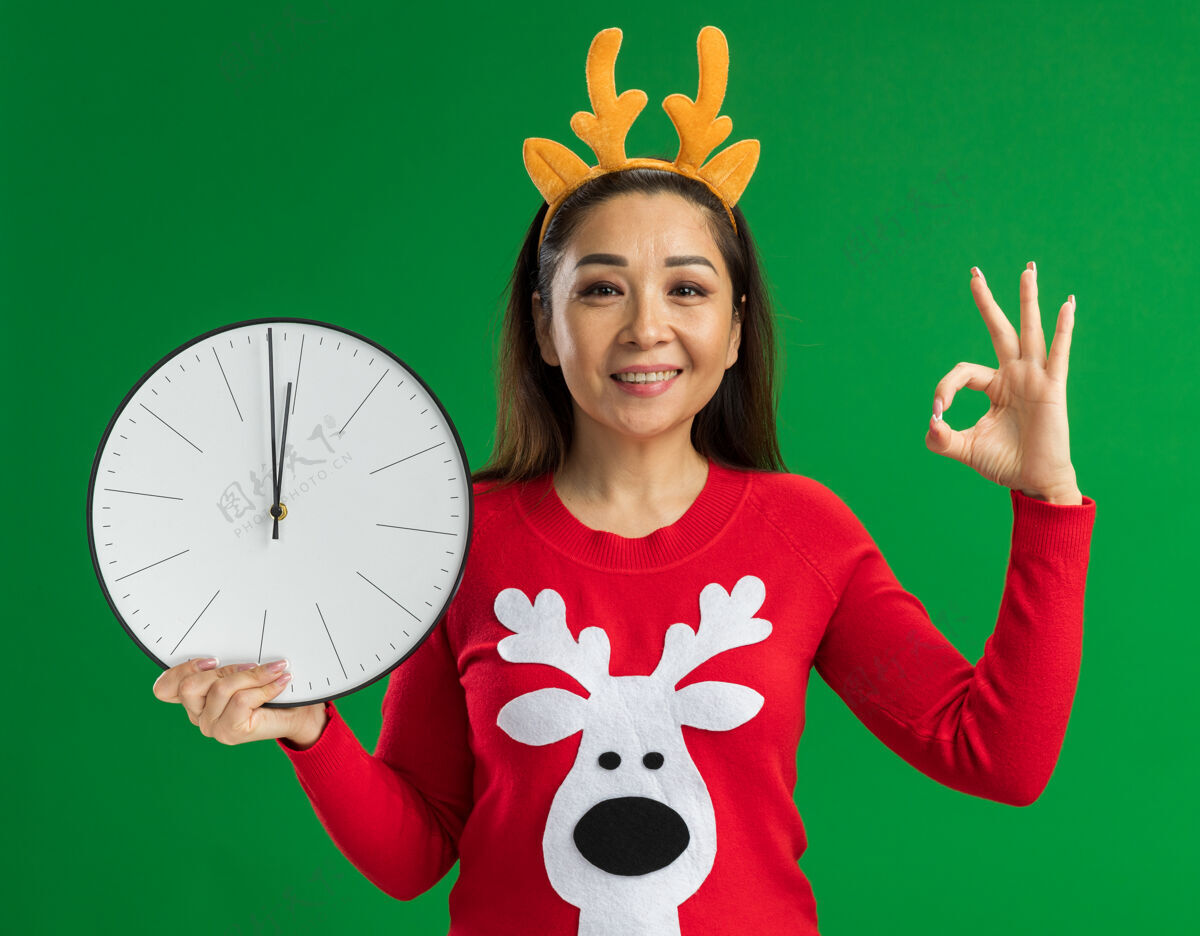 毛衣戴着鹿角和红色毛衣的圣诞戒指的年轻女子拿着挂钟微笑着看着相机 绿色背景上立着ok标志角钟站着