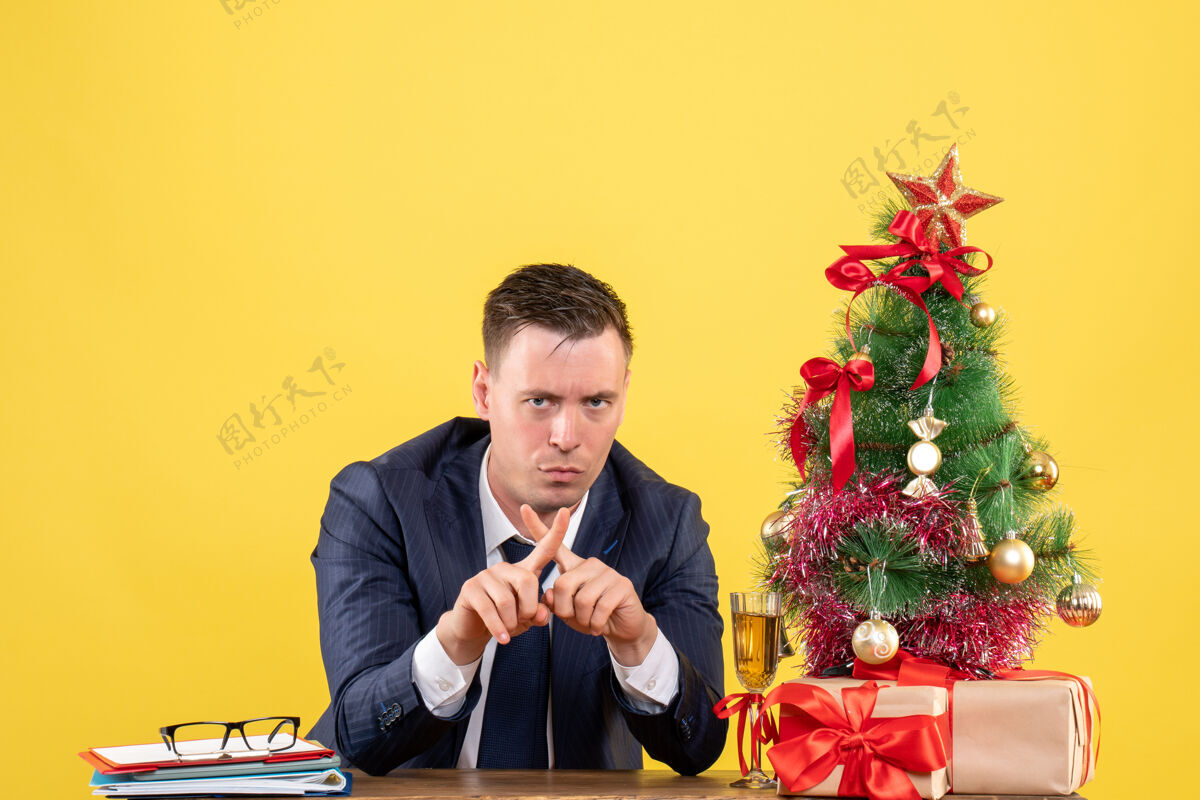 树不满意的男人交叉手指坐在圣诞树旁的桌子前 黄色的礼物坐着交叉新郎