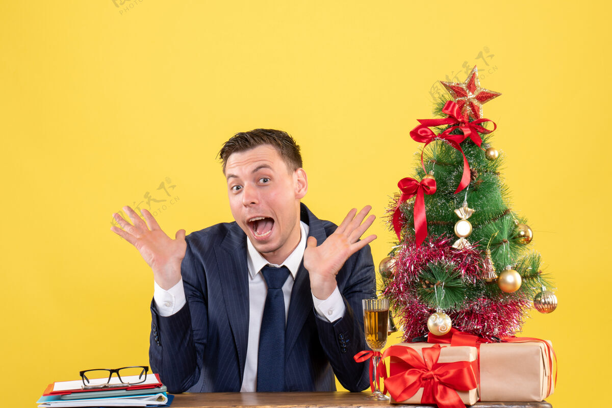 开口前视图惊讶的人张开双手张开嘴坐在圣诞树和礼物旁边的桌子上男人桌子嘴巴