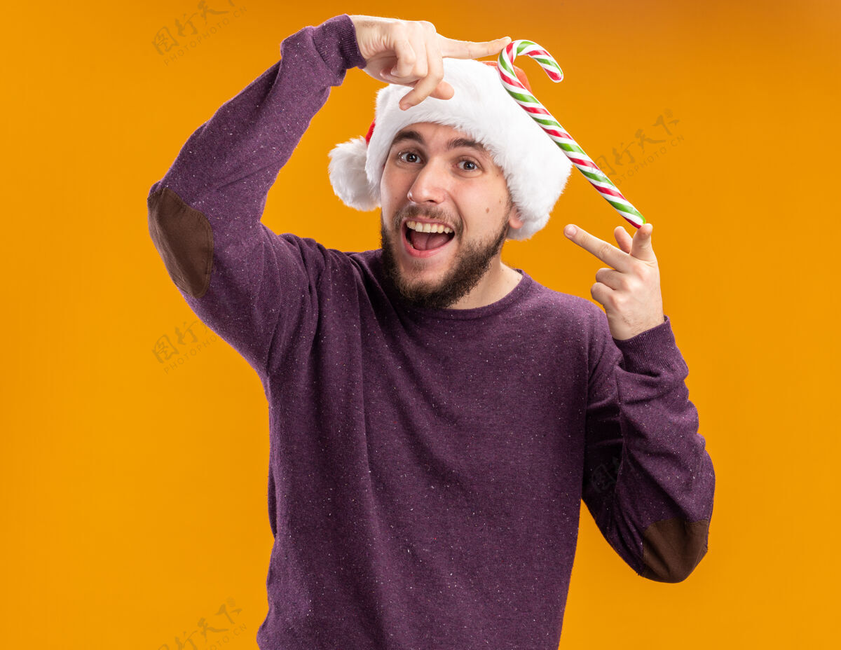 圣诞节身穿紫色毛衣 戴圣诞帽 手持糖果棒的年轻人站在橘色的墙上开心地微笑着糖果站着毛衣
