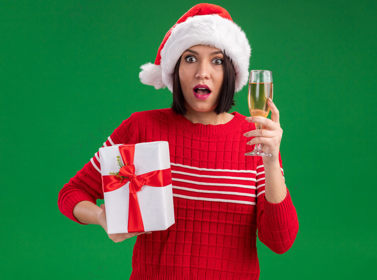 香槟给人印象深刻的年轻女孩戴着圣诞帽拿着礼包和一杯香槟隔离在绿色的墙上圣诞老人包装玻璃