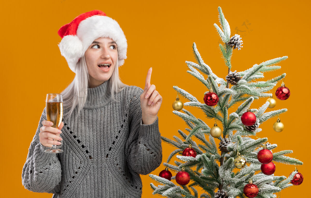 圣诞年轻的金发女郎穿着冬天的灰色毛衣 戴着圣诞帽 手里拿着一杯香槟 食指朝上 快乐而愉快地站在橙色背景下的圣诞树旁索引圣诞树圣诞快乐
