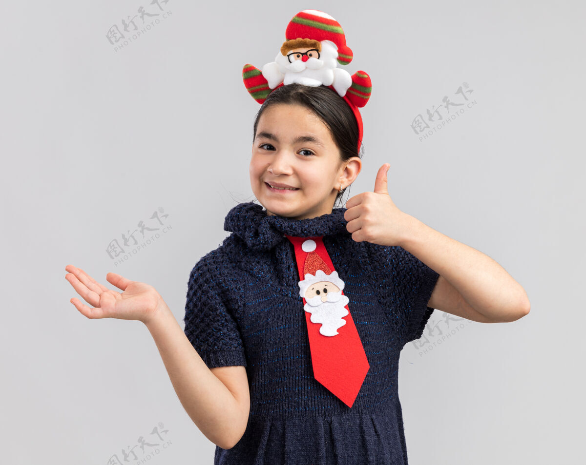 站着小女孩穿着针织连衣裙 头上系着一条红色领带 头上戴着一个有趣的圣诞戒指 面带微笑 竖起大拇指 用手臂展示复制空间向上红色空间