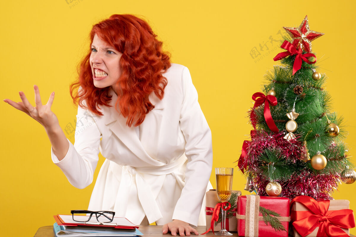 女医生前视图：女医生坐在桌子后面 拿着圣诞礼物 在黄色背景上争论着圣诞树和礼品盒情感微笑礼物
