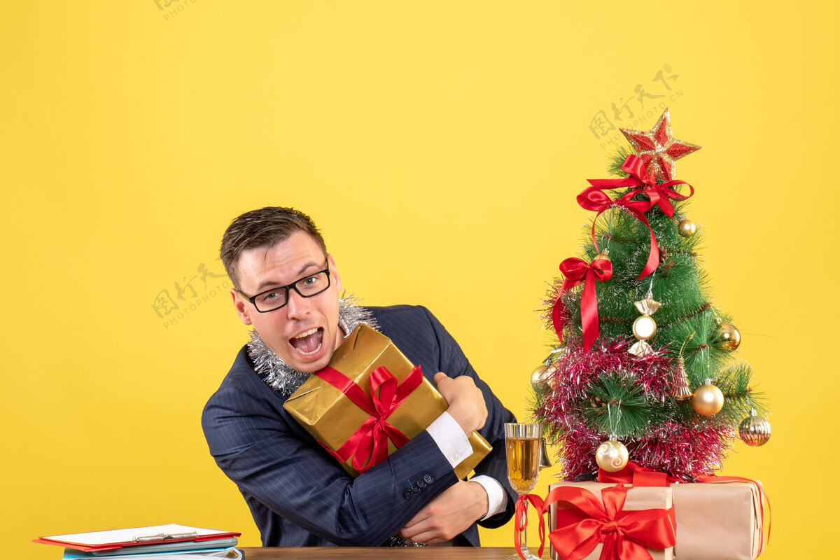 抱着正面图：圣诞树旁的桌子上 快乐的男人紧紧地拿着礼物 黄色的礼物圣诞节树快乐的男人