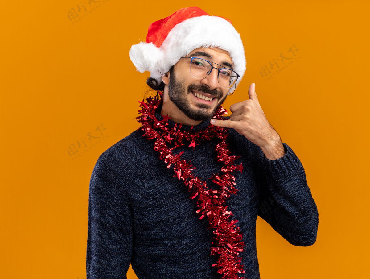 帅气带着微笑的年轻帅哥戴着圣诞帽 脖子上戴着花环 在橙色背景下显示出孤立的打电话手势手势电话花环