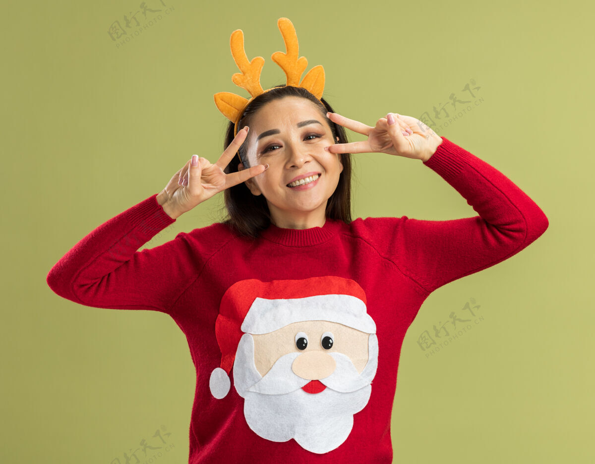 鹿穿着红色圣诞毛衣的年轻女子 戴着滑稽的鹿角边 看着摄像机 脸上带着幸福的笑容 眼睛旁边有个v字 站在绿色的背景上眼睛脸Vsign