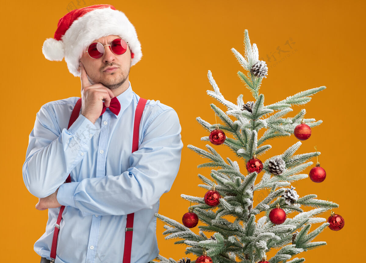 年轻戴着圣诞帽 戴着红眼镜 系着吊带领结的年轻人站在圣诞树旁 脸上带着沉思的表情 想着橙色的墙蝴蝶结思想男人