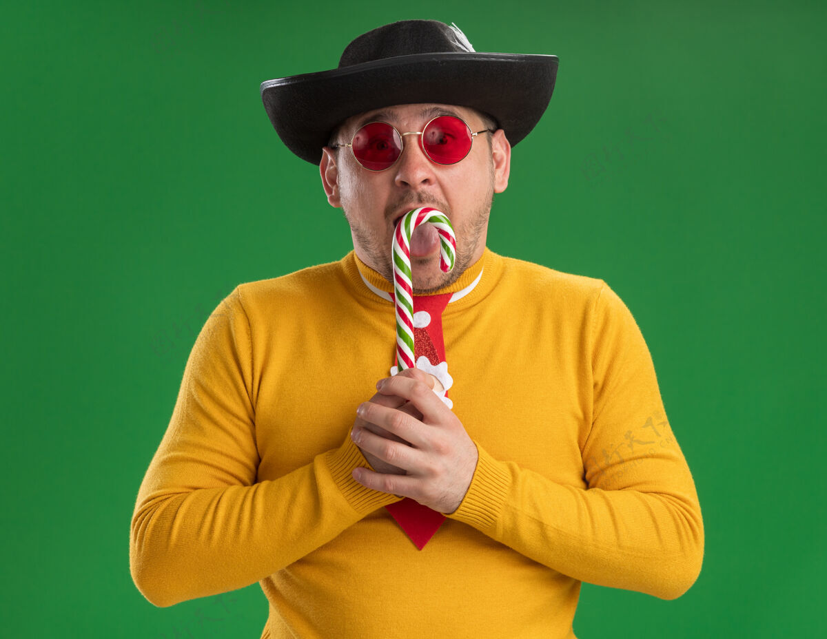 站着一个穿着黄色高领毛衣 戴着滑稽的红领带 戴着黑色帽子 手里拿着糖果棒的年轻人站在绿色的墙壁上惊讶不已圣诞节领带年轻