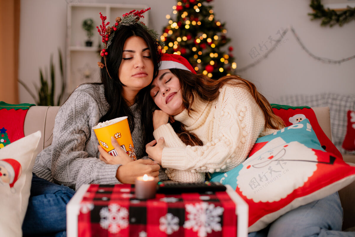 家带着圣诞帽和冬青花环的困倦漂亮的年轻女孩抱着爆米花桶坐在扶手椅上 在家里享受圣诞节时光快乐爆米花享受