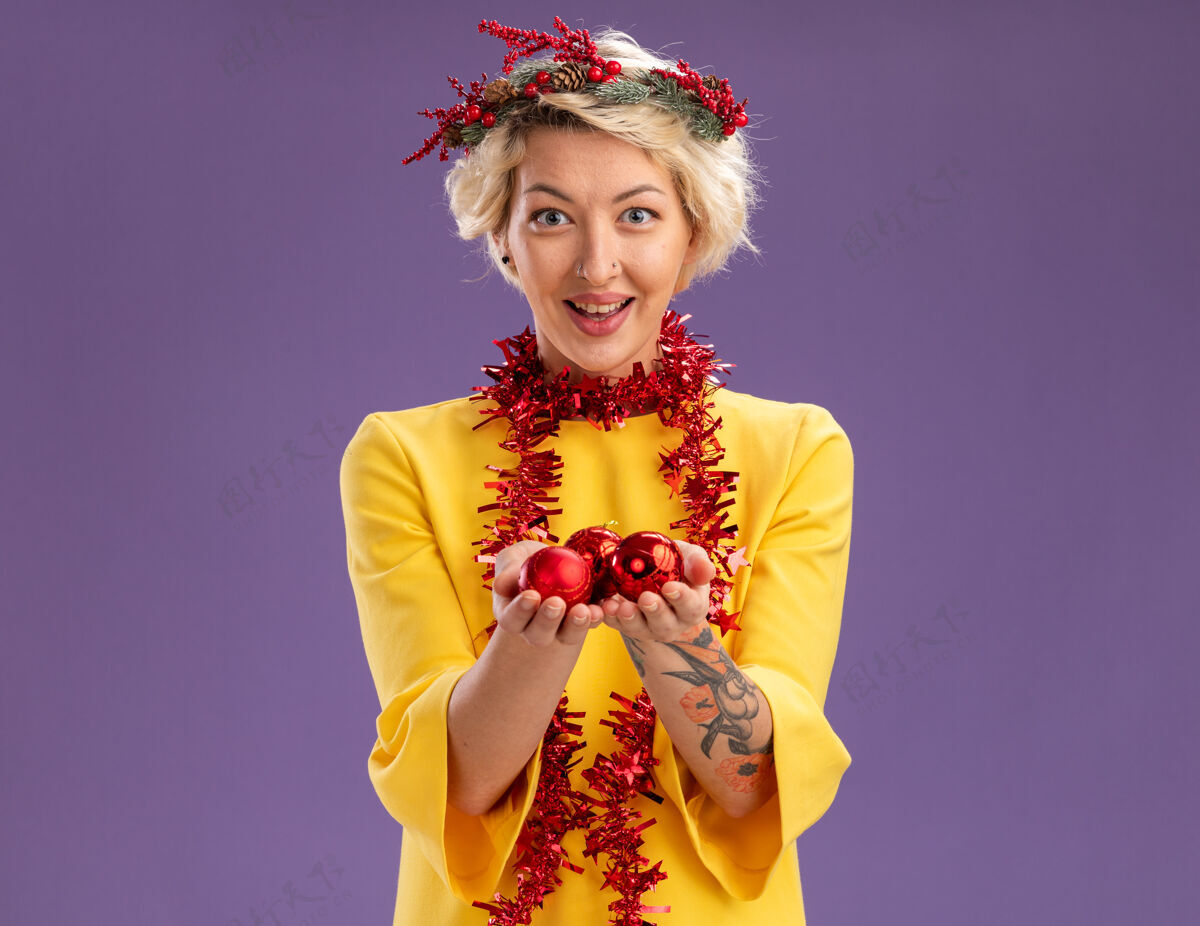 花环令人印象深刻的年轻金发女子头戴圣诞花环和金箔花环在脖子上看持有圣诞饰品孤立在紫色墙上复制空间金属丝圣诞节脖子