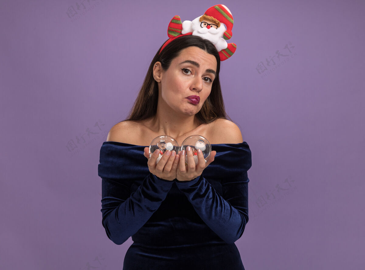 舞会迷茫的年轻漂亮女孩穿着蓝色的裙子 打着圣诞发箍 手里拿着紫色背景上孤立的圣诞球女孩困惑抱着