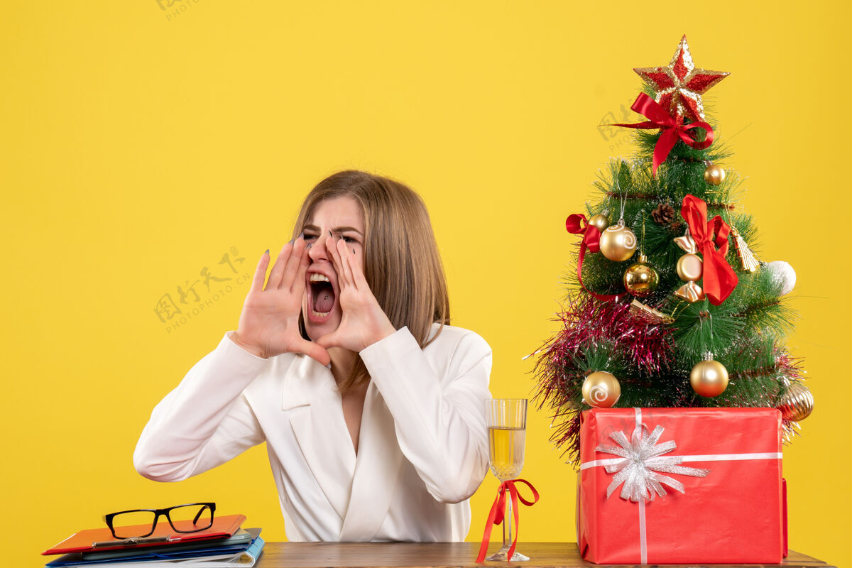 新的前视图女医生坐在她的桌子前大声呼唤黄色背景与圣诞树和礼品盒桌子女商人呼叫