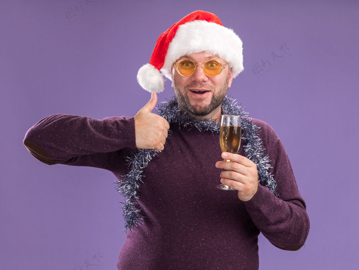 金属片令人印象深刻的中年男子戴着圣诞老人帽 脖子上戴着金箔花环 戴着一副眼镜 手里拿着一杯香槟 在紫色的墙上孤立地竖起大拇指拇指中年帽子