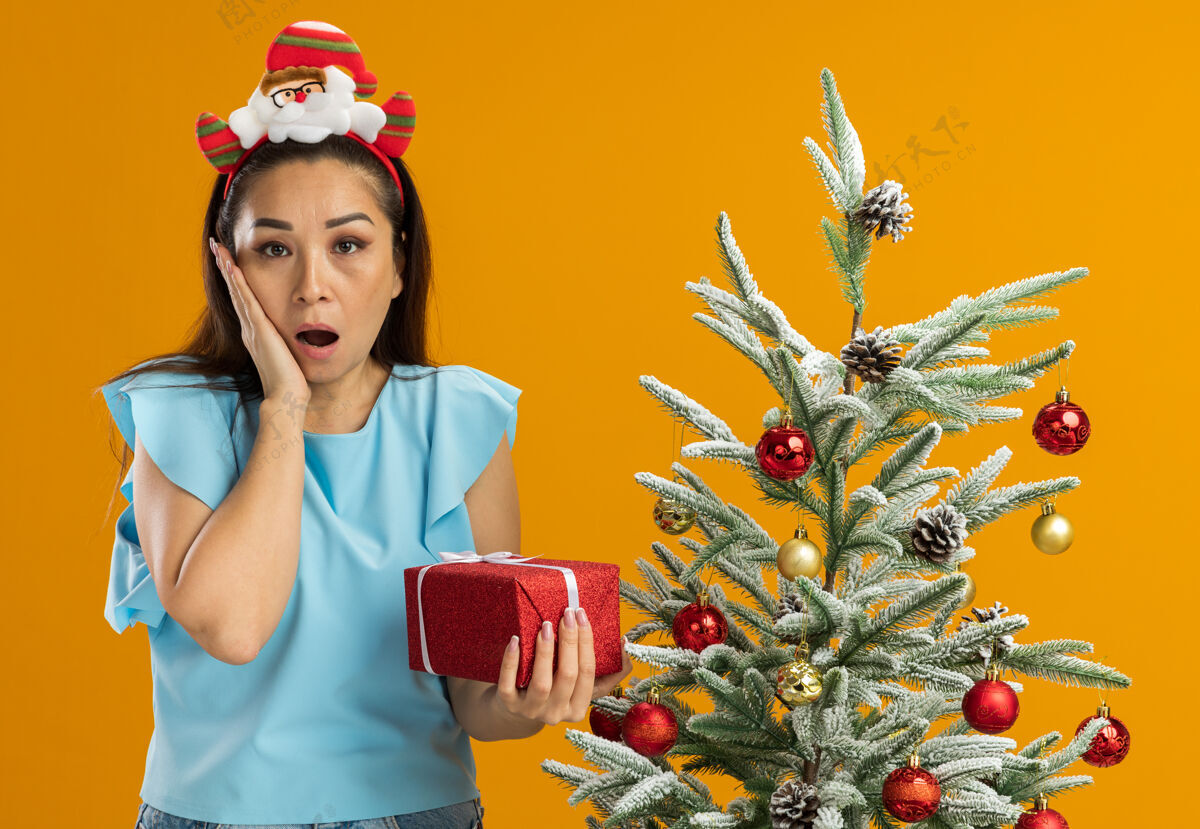 树身穿蓝色上衣的年轻女子头上戴着有趣的圣诞戒指 手里拿着礼物看着相机 手放在脸颊上 站在橙色背景下的圣诞树旁 惊讶不已手圣诞橘子