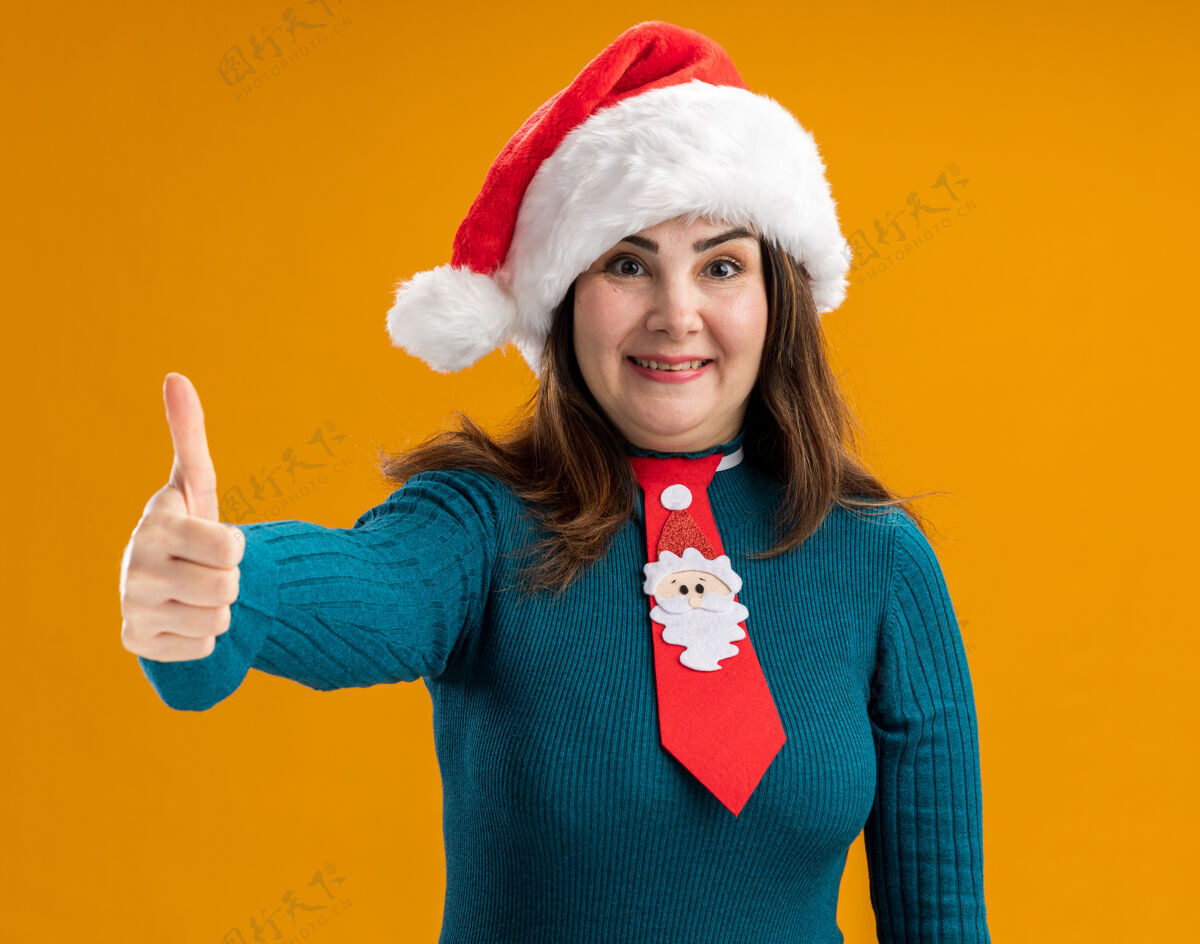 成人印象深刻的成年白人妇女与圣诞老人帽子和圣诞老人领带大拇指向上孤立的橙色背景与复制空间圣诞老人领带帽子