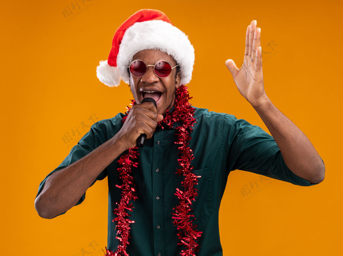 圣诞节戴着圣诞帽 戴着花环 戴着眼镜的非裔美国人举起手臂站在橙色的墙上对着麦克风喊叫非洲人举起来喊着