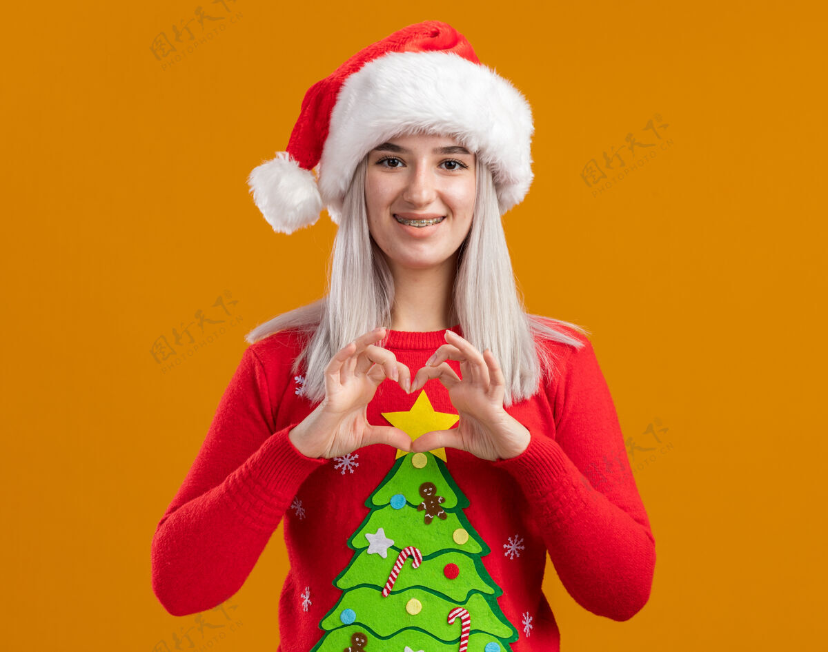 做身穿圣诞毛衣 戴圣诞帽的金发女郎面带微笑地看着摄像机 用手指在橙色背景上做着心形手势手指心年轻