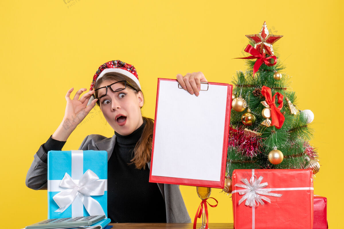 女孩正面图戴着圣诞帽的大眼睛女孩坐在桌旁摘下眼镜圣诞树和礼物鸡尾酒纸装饰礼物