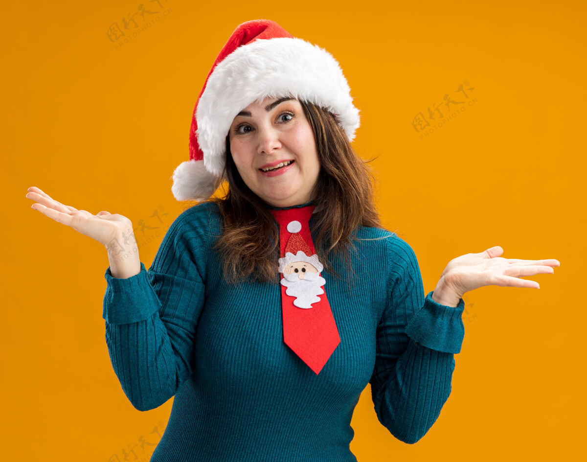 打开高兴的成年白人妇女戴着圣诞帽 打着圣诞领带 双手张开 孤立地放在橙色背景上 留有复制空间拿着帽子高兴