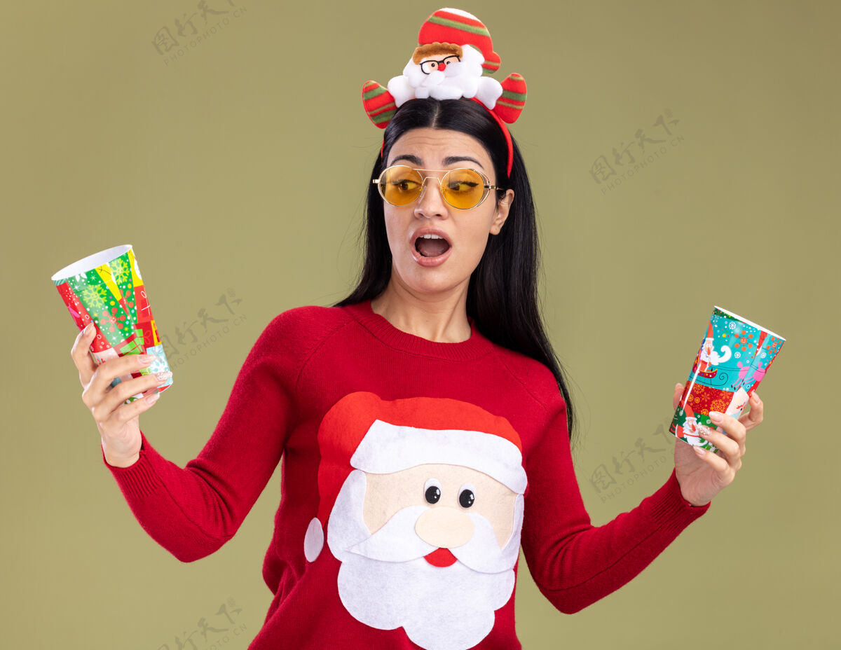 橄榄印象深刻的年轻白人女孩戴着圣诞老人的头带和毛衣 戴着眼镜 举着塑料圣诞杯 看着其中一个孤立在橄榄绿墙上塑料穿着圣诞老人