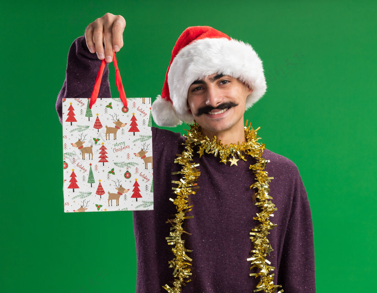 帽子快乐的小胡子男人戴着圣诞老人的帽子 脖子上戴着金属丝 展示着圣诞礼物的纸袋 站在绿色的背景下 面带微笑地看着相机脸金属片圣诞节