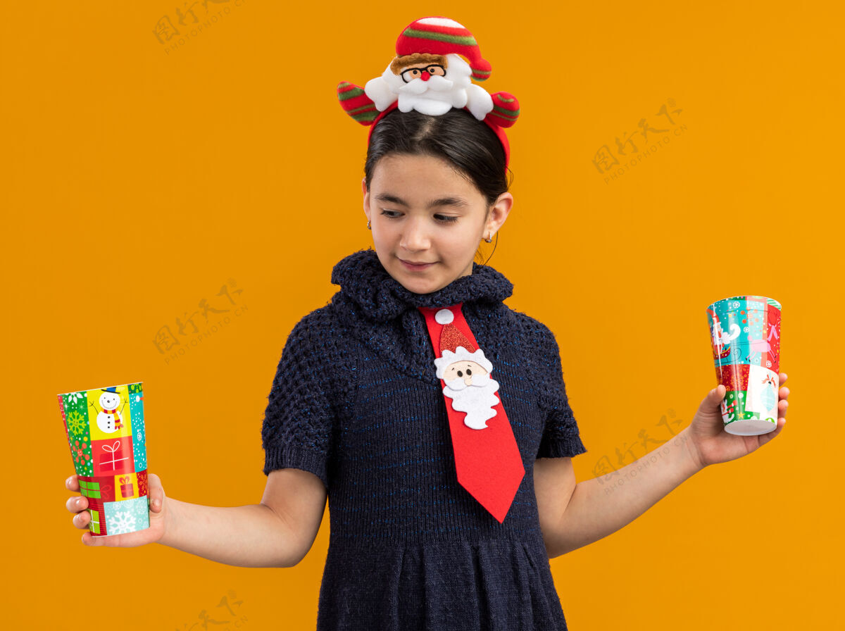 头穿着针织连衣裙的小女孩头上系着一条红色领带 手里拿着五颜六色的纸杯 站在橙色的背景上 看起来很困惑 试图做出选择编织选择边缘