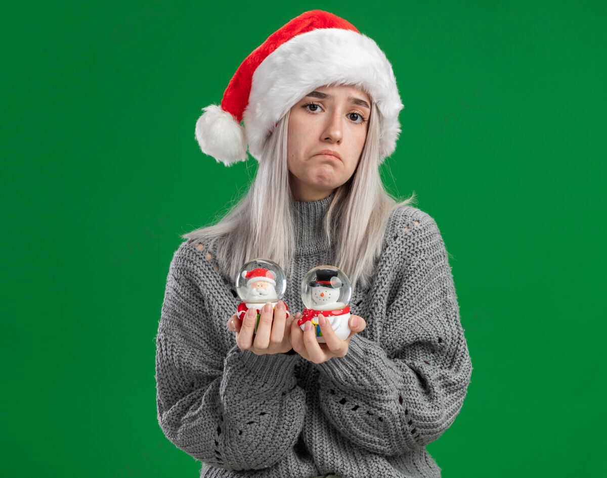 圣诞节年轻的金发女郎穿着冬衣 戴着圣诞帽 手里拿着圣诞玩具雪球 站在绿色的背景上困惑地看着摄像机 没有回答年轻金发回答