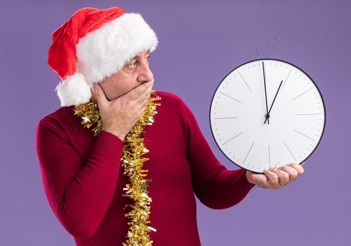 时钟中年男子戴着圣诞老人的帽子 脖子上戴着金属丝 手里拿着时钟看着它 忧心忡忡地捂着嘴 手站在紫色的背景上金属片周围站立