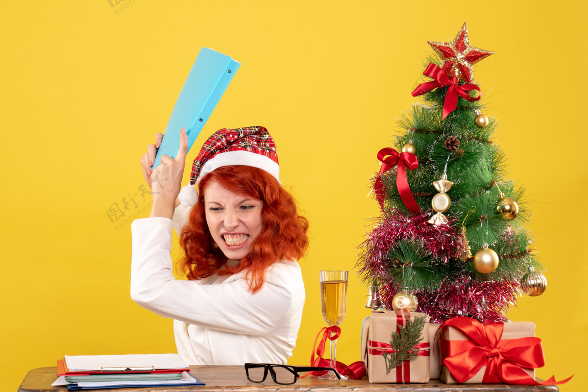 背景前视图：女医生坐在桌子后面 手里拿着文件 怒气冲冲地站在黄色背景上 手里拿着圣诞树和礼盒圣诞节快乐健康