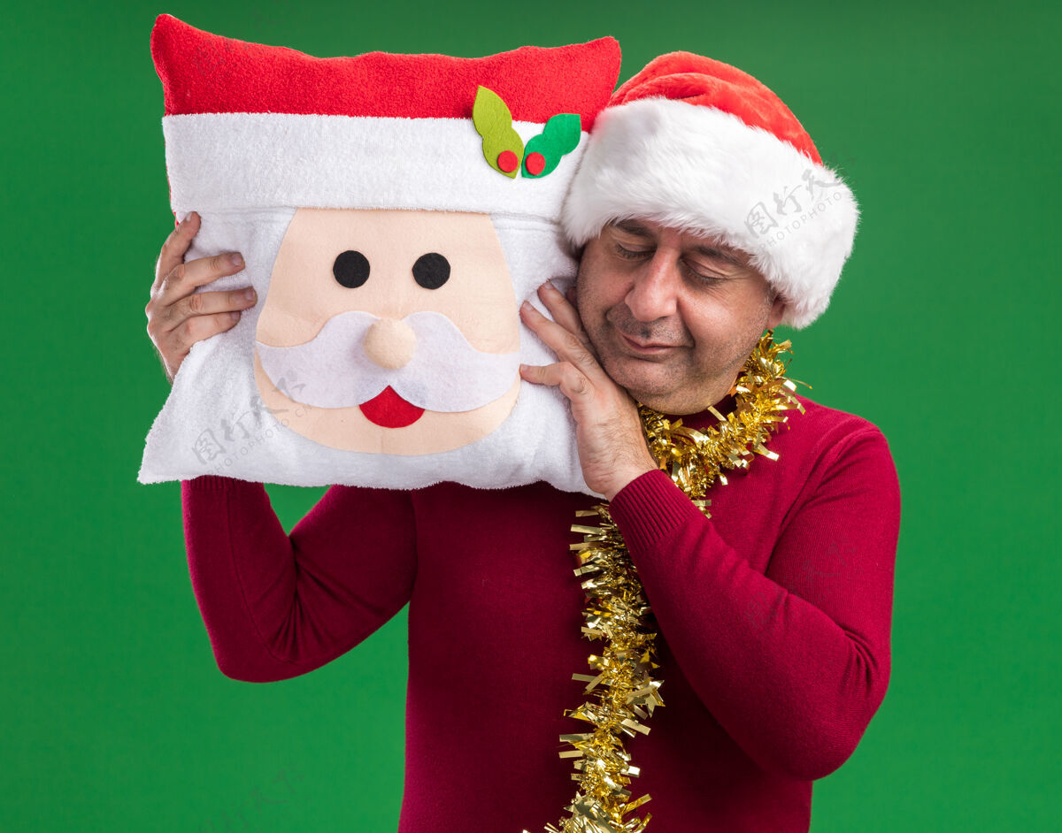 年龄中年男子戴着圣诞老人帽 脖子上戴着金属丝 抱着圣诞枕头 闭着眼睛站在绿色的背景上帽子金属片圣诞节