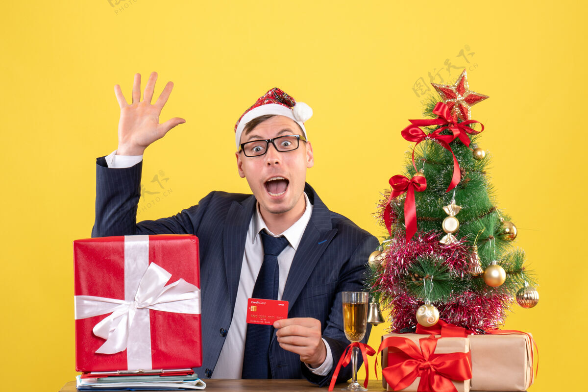 职业商务人士招呼某人坐在圣诞树旁的桌子前 并在黄色上展示礼物的正面视图桌子公司坐着