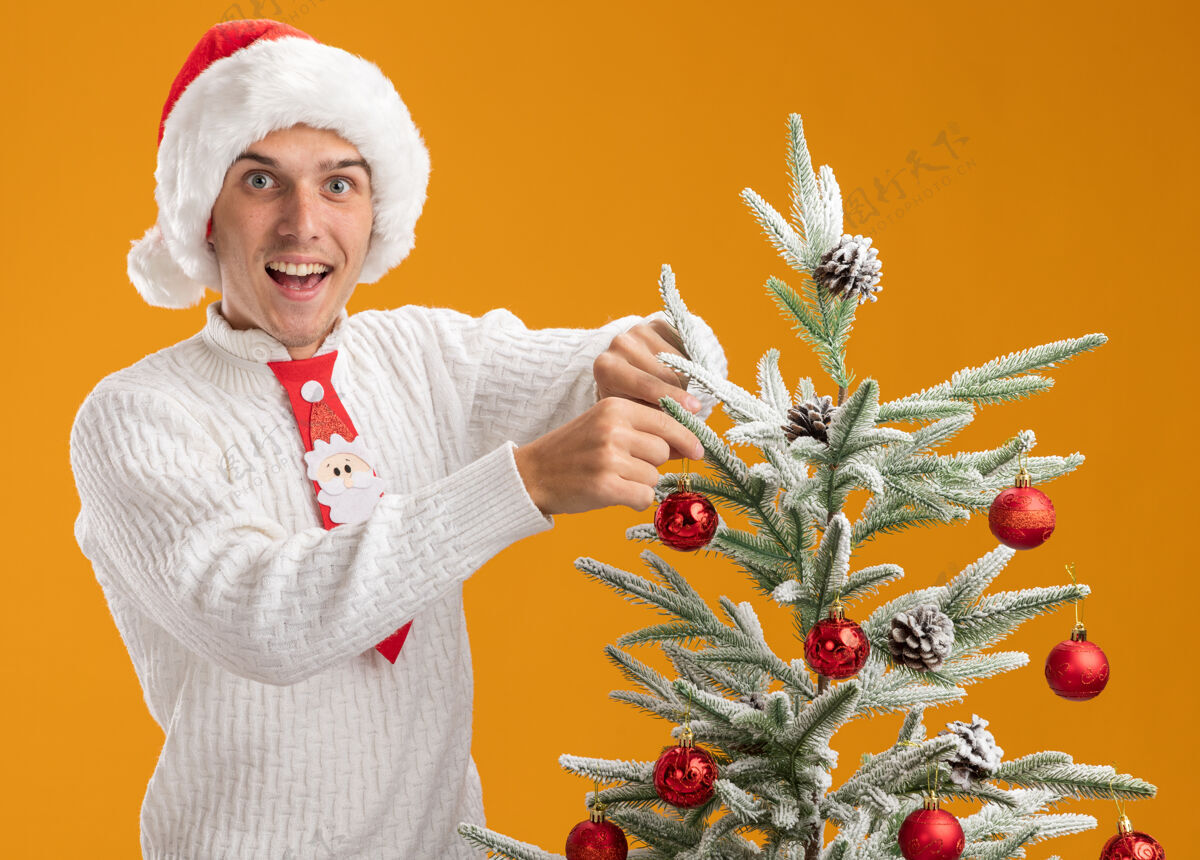 站着令人印象深刻的年轻帅哥戴着圣诞帽和圣诞老人领带站在圣诞树附近装饰圣诞球装饰隔离在橙色的墙上近年轻球